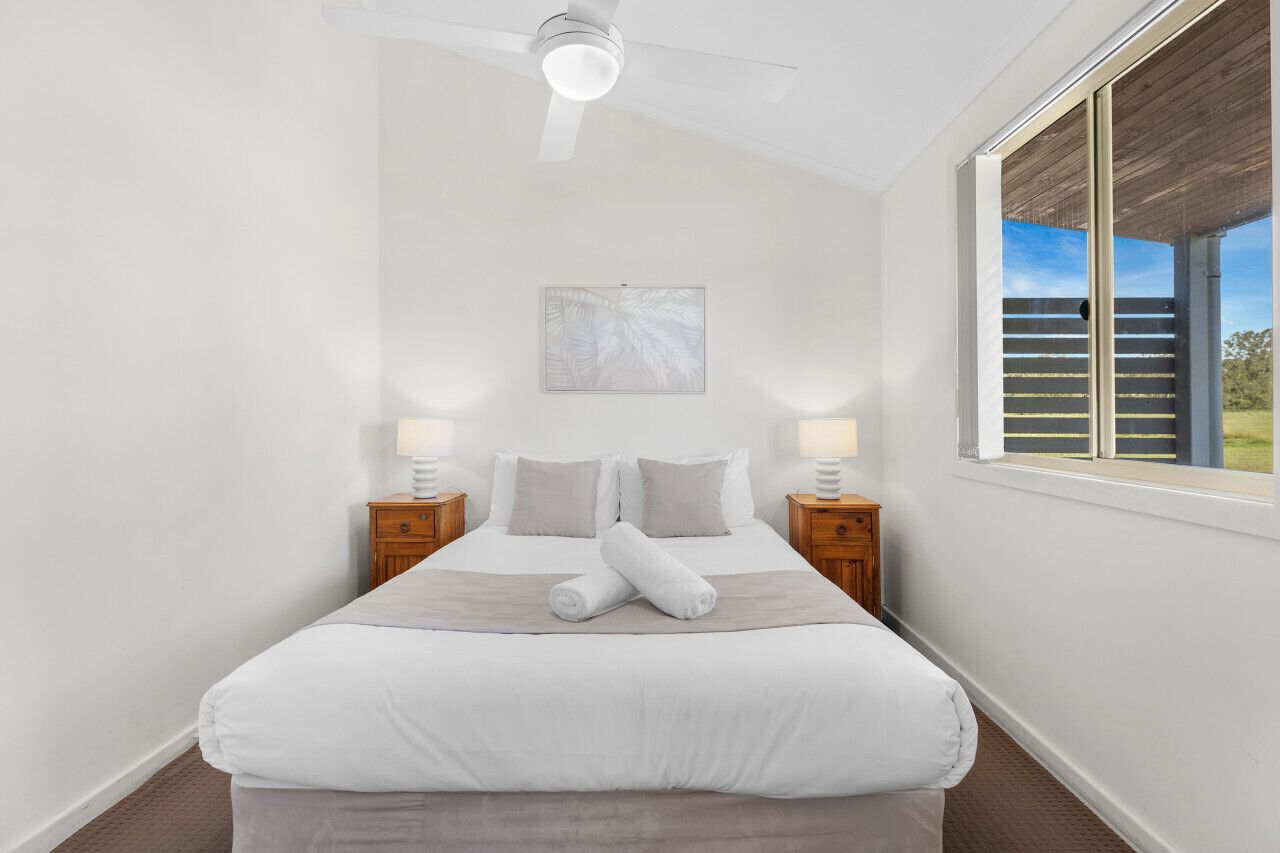 A neatly made bed with white linens and a gray throw, two bedside lamps, a ceiling fan, and a window with a view of a wooden deck.