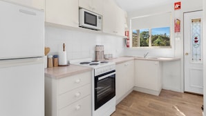 Una cocina moderna con armarios blancos, microondas, estufa y horno. Se ve una ventana con vistas a árboles y una puerta con letrero de salida de emergencia en rojo.