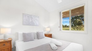 Una cama bien hecha con un cubrecolcha gris, almohadas blancas y un cabecero blanco. Hay una mesita de noche de madera con una lámpara, y una ventana con vistas a árboles y césped.