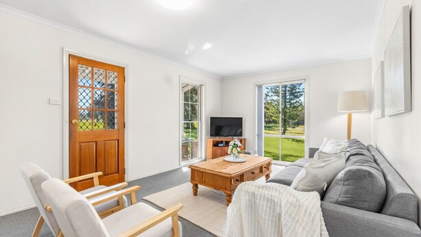 A living room with a grey sofa, a wooden coffee table, and a TV. There is a wooden door with a glass panel and a view of a green landscape through the windows. - Madigan Cottages | Settlers Cottage (Lovedale)