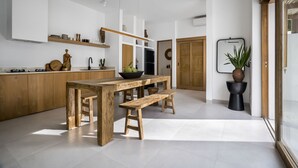 A modern kitchen with a wooden table and bench, white cabinetry, and a potted plant. - Villa Oasi 2 by Alfred in Bali (Pecatu)