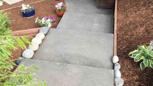 Un patio couvert aménagé avec des meubles en osier, des plantes en pot et un chemin en béton.