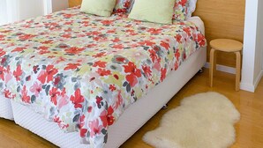 Ein Schlafzimmer mit einem Holzkopfteil, einer blumigen Tagesdecke, zwei Kissen, einem kleinen Holzschemel, einem flauschigen Teppich und einer Wandleuchte.