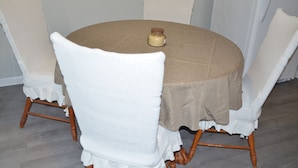 Mesa redonda com toalha de mesa bege, rodeada por quatro cadeiras brancas de bordas franjadas.