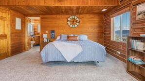 A log cabin bedroom with a bed, a clock on the wall, a bookshelf, and a window with a view.