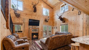 A log cabin interior with a fireplace, wooden walls, mounted deer heads, and a cozy seating area.