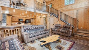 A spacious log cabin interior with a living area featuring two sofas and a coffee table, a kitchen with bar stools, and a staircase leading to an upper level.