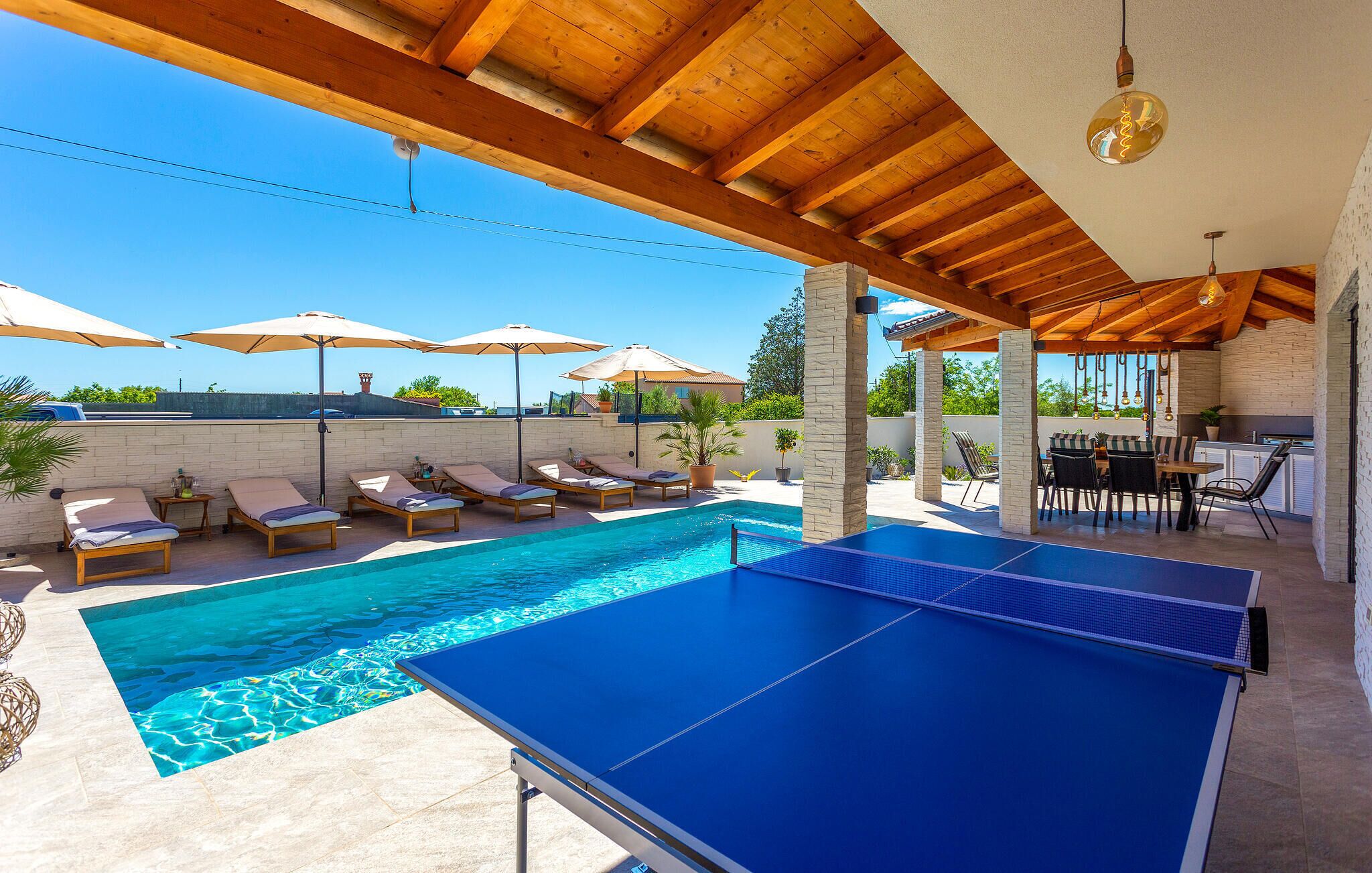 A pool area with lounge chairs and umbrellas, a ping pong table, and a barbecue area.