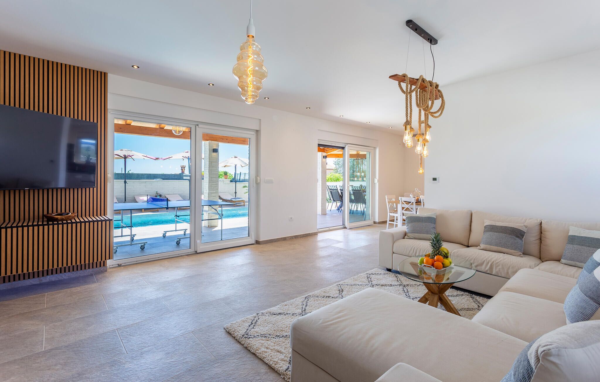 A modern living room with a flat-screen TV, a sofa set, a glass coffee table, and a pool area visible through glass doors.