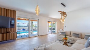 Ein modernes Wohnzimmer mit Flachbildfernseher, einer Sofagarnitur, einem Glastisch und Blick auf einen Pool durch Glasüren.