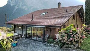 Eine Chalet mit rotem Ziegeldach, einer gläsernen Veranda und einem Garten mit Hortensien.