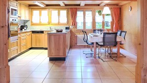 Eine Holzdiele mit Küche, die eine zentrale Kochinsel, einen Essbereich mit Tisch und Stühlen sowie große Fenster mit Vorhängen umfasst.