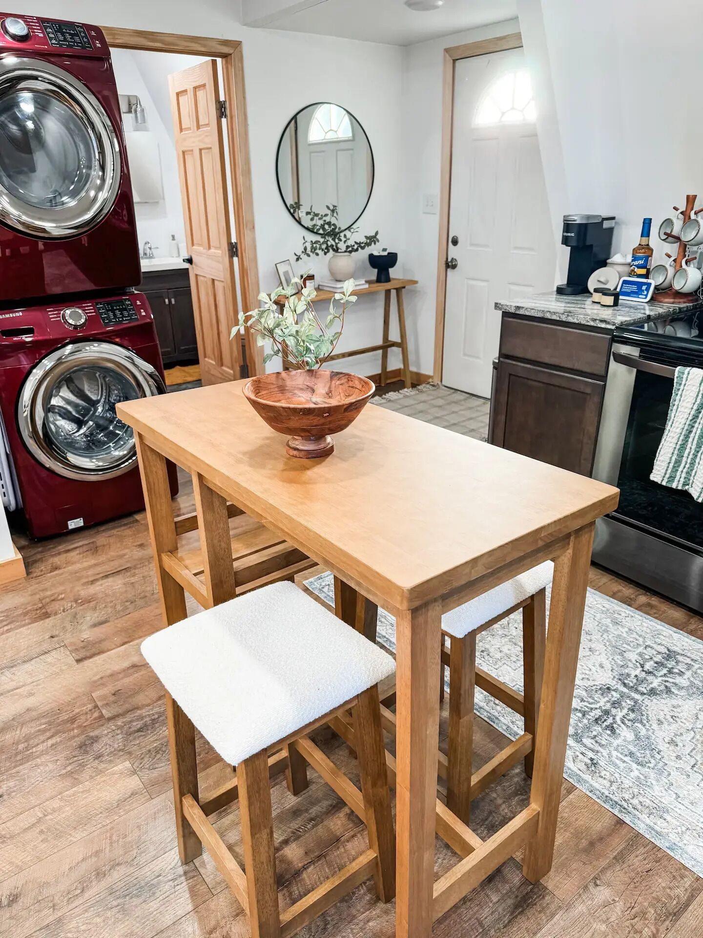 Une cuisine compacte avec une table et un banc en bois, un lave-linge rouge, un miroir et un petit plan de travail sur lequel sont posés des ustensiles de cuisine.