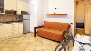 A compact kitchen with a stove, refrigerator, and white cabinets. A wooden chair and a small table with a plate are visible. - Casa Bartolomea - CASA BARTOLOMEA 11 (Marina di Campo)