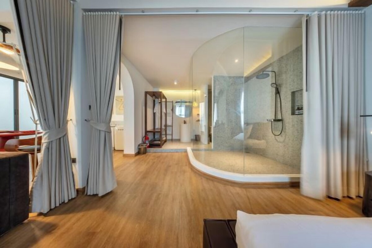 A modern hotel room with a curved glass shower enclosure, wooden flooring, and a bed with white bedding.