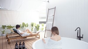 A modern outdoor relaxation area with a wooden deck, a lounge chair, a bathtub, and potted plants. - The Lair Bangkok (Bangkok)