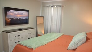 Ett sovrum med en säng, ett orange överkast, en grön pläd, en vit kudde, ett nattduksbord, en spegel, en tv på väggen och ett fönster med vita gardiner.