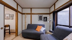 A compact room with a sofa, a chair, and a small table. There are framed pictures on the wall, a window with curtains, and a wooden floor.
