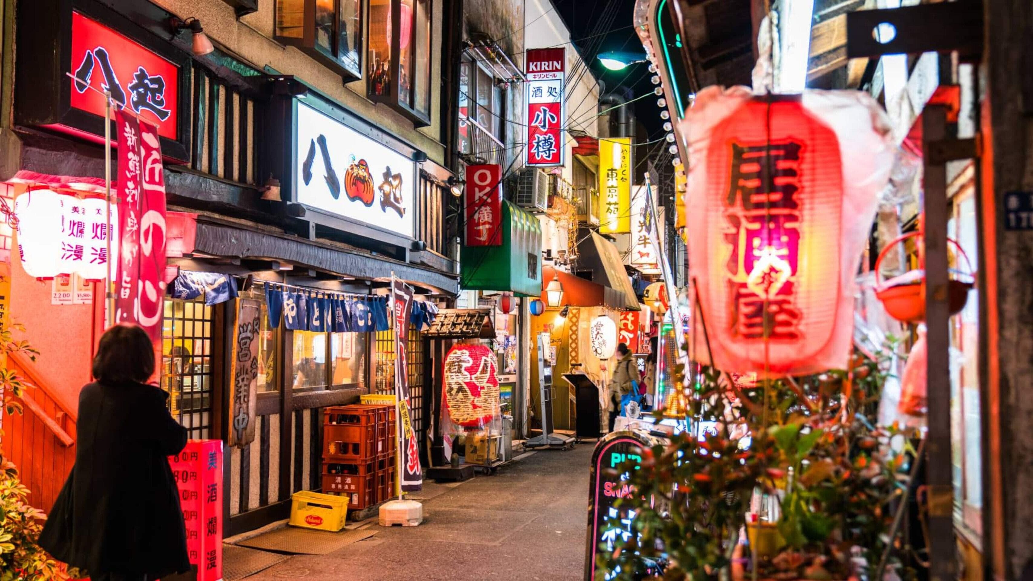 A narrow street lined with shops and restaurants, featuring illuminated signs and storefronts.