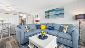 Un salon avec un canapé d’angle bleu, une table basse blanche et un tableau représentant l’océan accroché au mur.