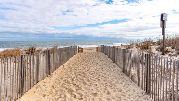 Un sentier de sable qui longe la plage et mène à l’océan, bordé d’une clôture en bois.