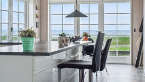 Een keuken met een groot raam dat uitkijkt op de zee, een groene plant op het aanrecht, en een zwarte stoel.