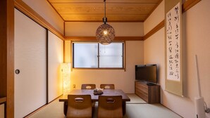 A traditional Japanese room with tatami flooring, a low wooden table with cushions, a television, and a hanging light fixture.