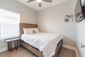 Un dormitorio con cama, ventilador de techo, ventana con persianas y una mesita.