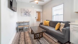 Ein Wohnzimmer mit Sofa, Couchtisch, Fernseher, Deckenventilator, Kunstwerken und einer Tür mit Jalousien.