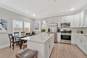 A modern kitchen with white cabinetry, a central island, a dining area with chairs, and a large window with blinds.