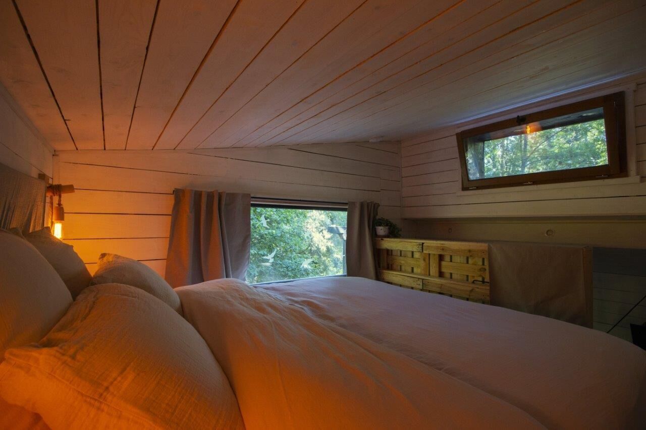 Ein gemütliches Schlafzimmer mit einem großen Bett, einem hölzernen Kopfteil, einem Fenster mit Vorhängen und Blick auf Bäume.