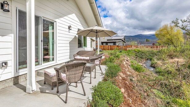 Ein Haus mit überdachter Terrasse, Korbmöbeln und Blick auf einen Bach und Berge.