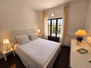 A neatly arranged hotel room with a large bed, two bedside tables with lamps, a small table with a book, and a window with curtains.