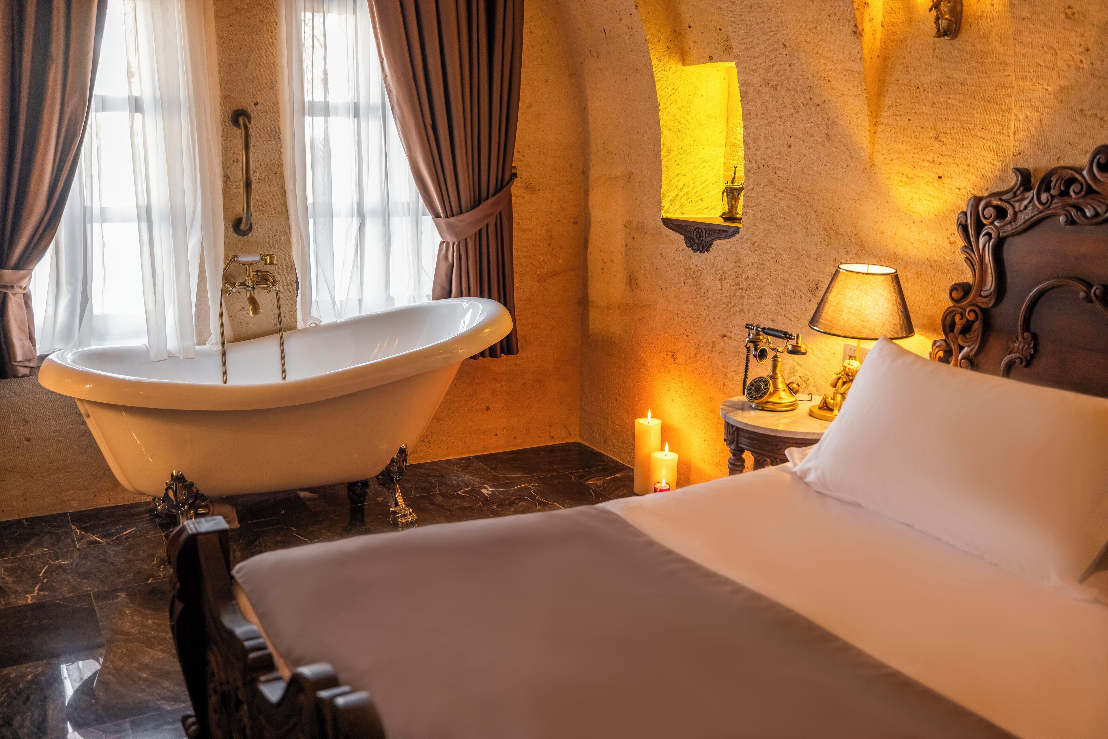 A room with a large bathtub, a bed with white linens, a nightstand with a lamp, and a wall-mounted mirror.