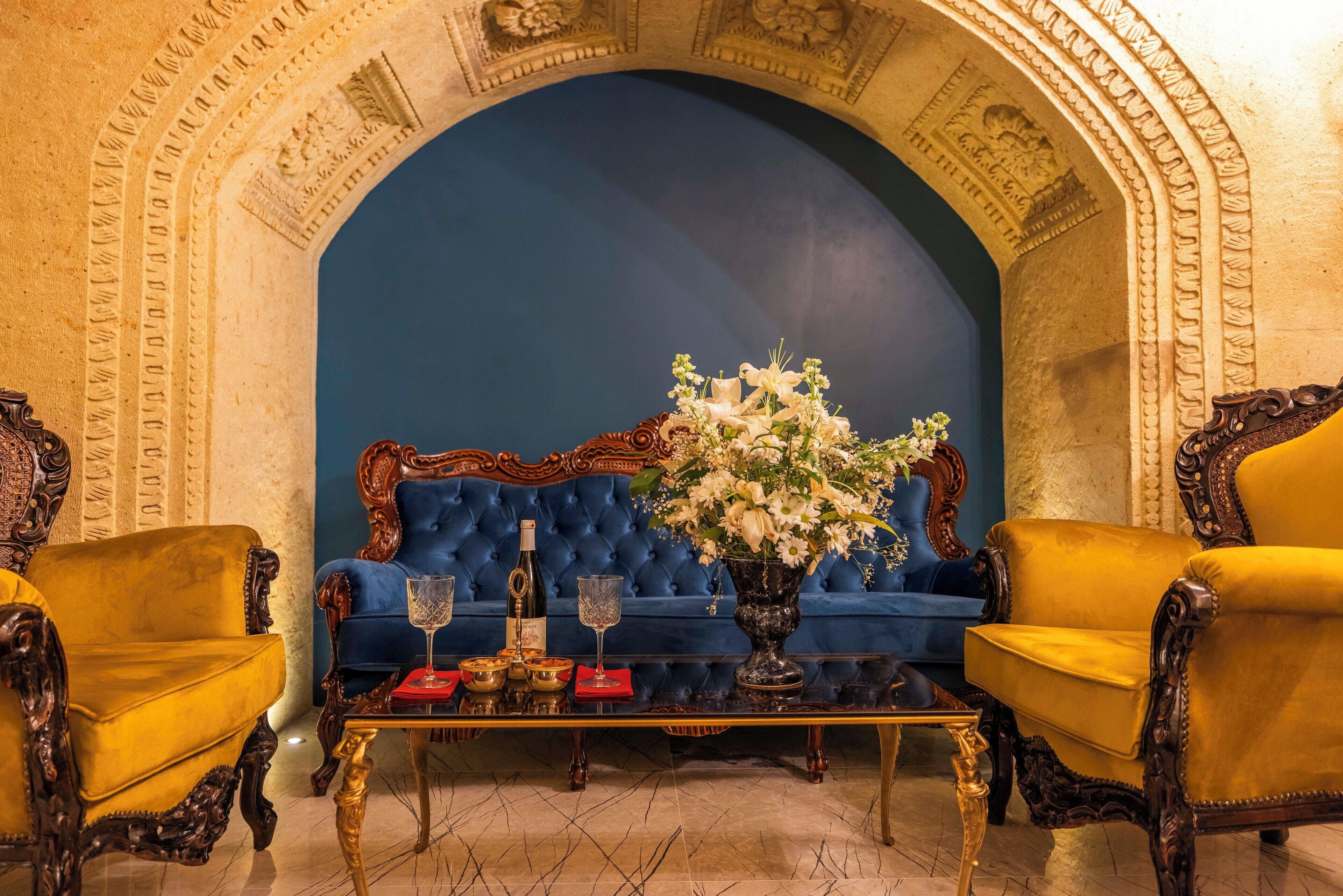 Une chambre luxueuse avec un canapé bleu recouvert de tissu, des fauteuils en bois richement décorés, une table basse en verre et un vase décoratif rempli de fleurs.
