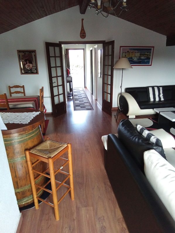 Un vaste espace de vie doté d’un plancher en bois, d’un canapé noir et blanc, d’une chaise en bois et d’une table à manger avec des chaises. Une porte donne accès à une autre pièce.
