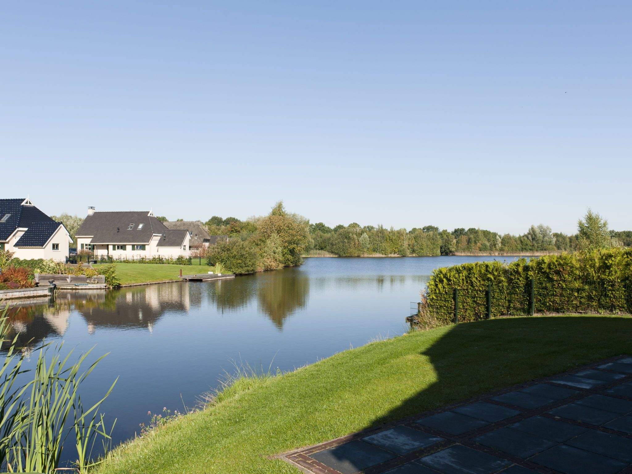 A serene lakeside view with a grassy area, a wooden dock, and houses in the background.