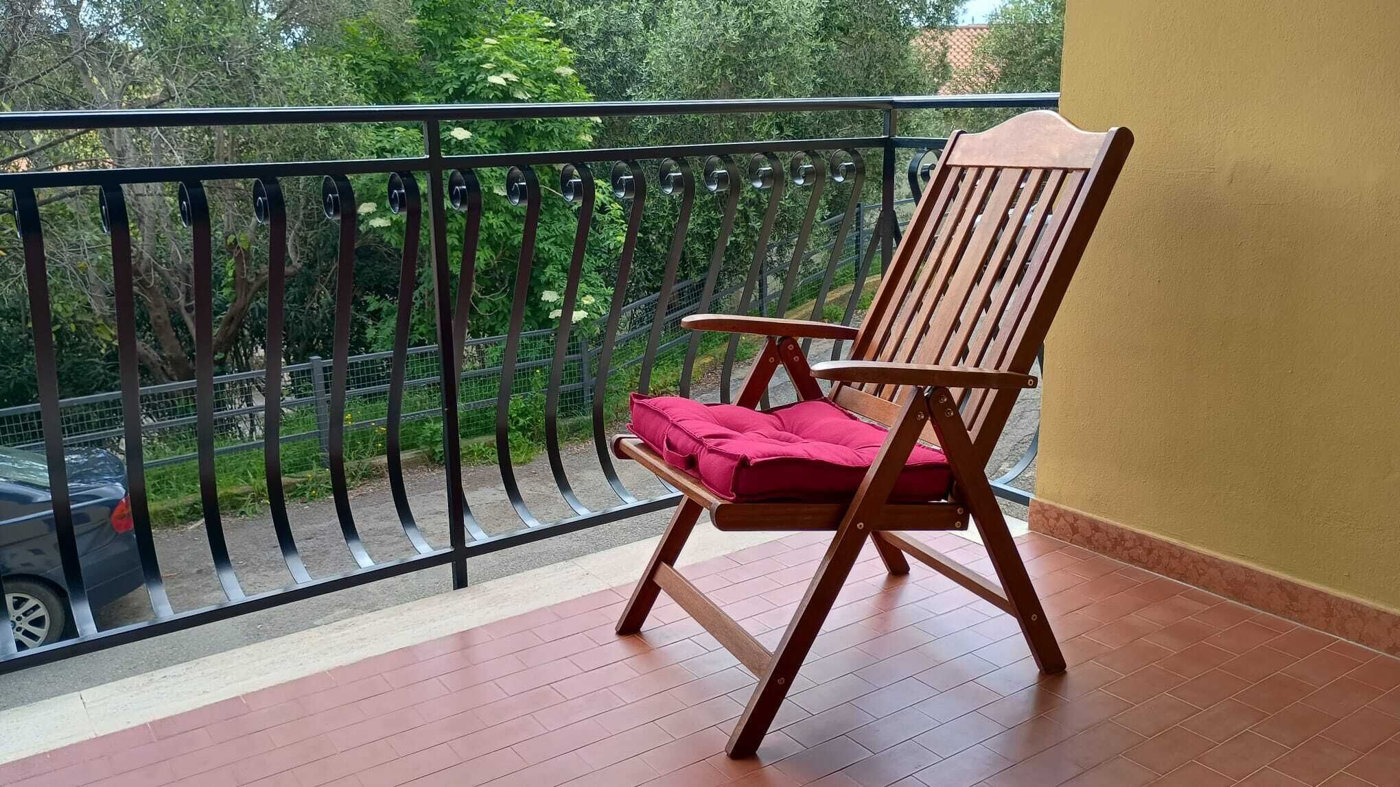 Ein Holzsessel mit rotem Kissen auf einem Balkon mit Metallgeländer, mit Blick auf eine Straße und Grünflächen.