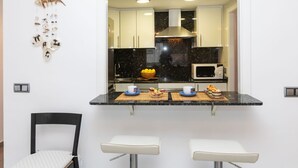 Una cocina moderna con armarios blancos, encimera negra y un microondas. Hay dos taburetes blancos y una pequeña mesa con un cuenco de limones y una taza.