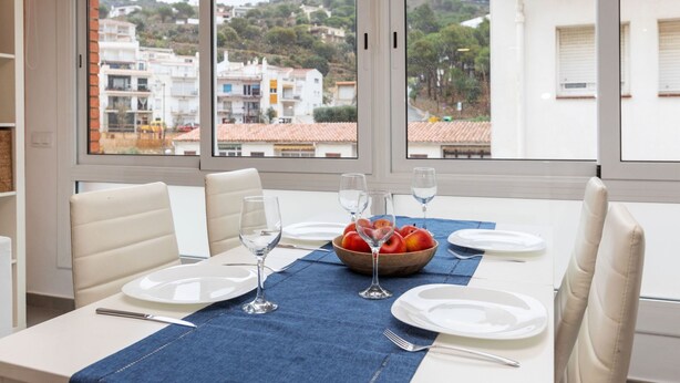 Un comedor con una mesa lista para dos personas, un frutero y copas de vino. Amplias ventanas ofrecen vistas a edificios y árboles.