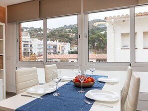 Un comedor con una mesa lista para dos personas, un frutero y copas de vino. Amplias ventanas ofrecen vistas a edificios y árboles.