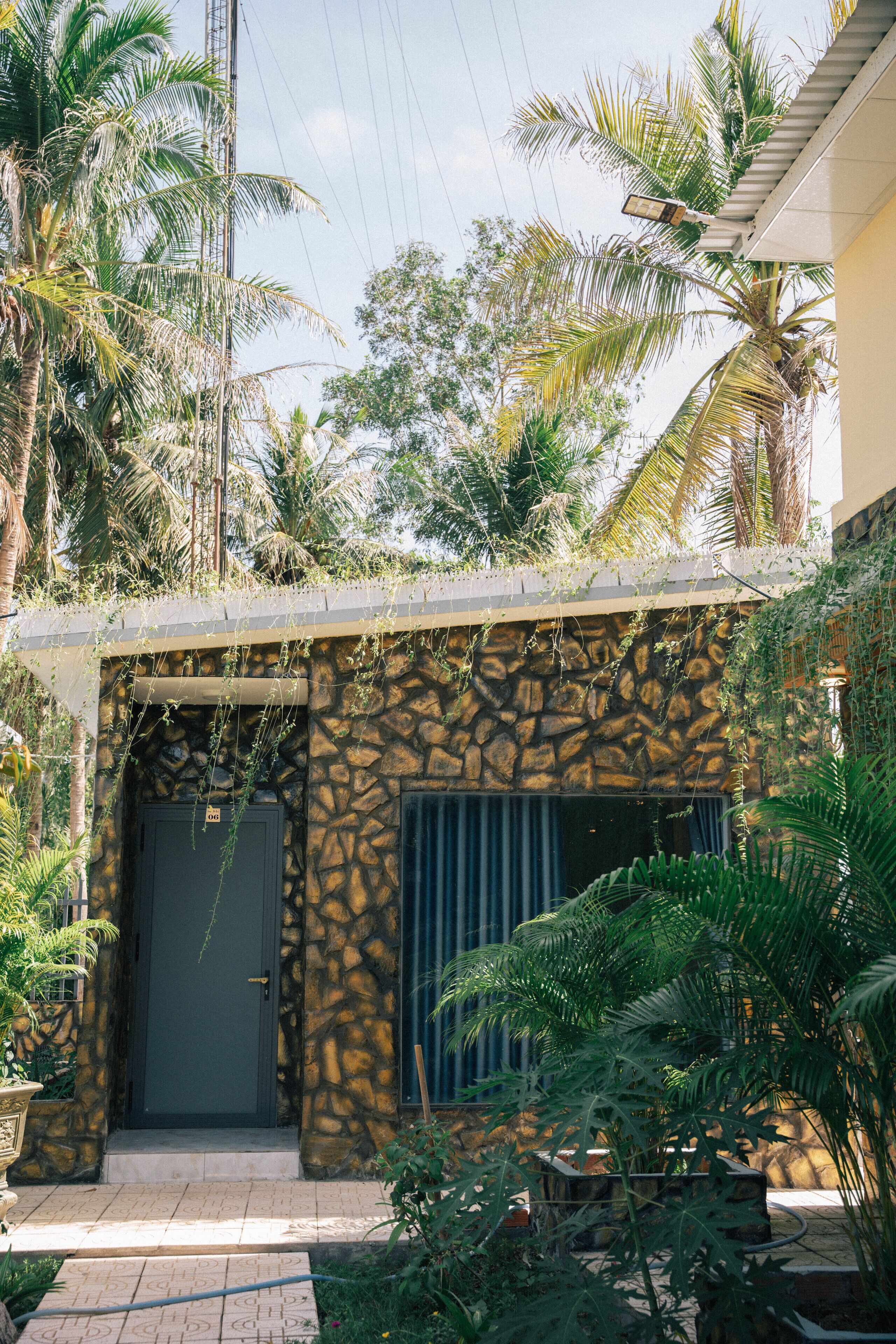 A building with a stone facade, a dark door, and a small garden with palm trees.