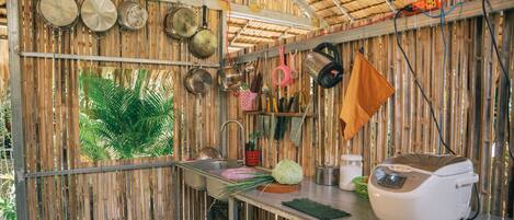 Una cocina con paredes de bambú, diferentes utensilios de cocina, un lavamanos y una cocedora de arroz.