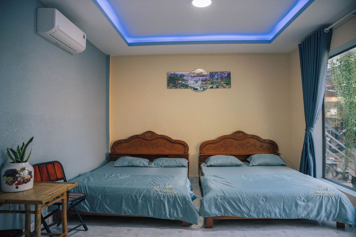 A room with two beds, a ceiling light, a wall-mounted air conditioner, a table with a potted plant, and a window with curtains.