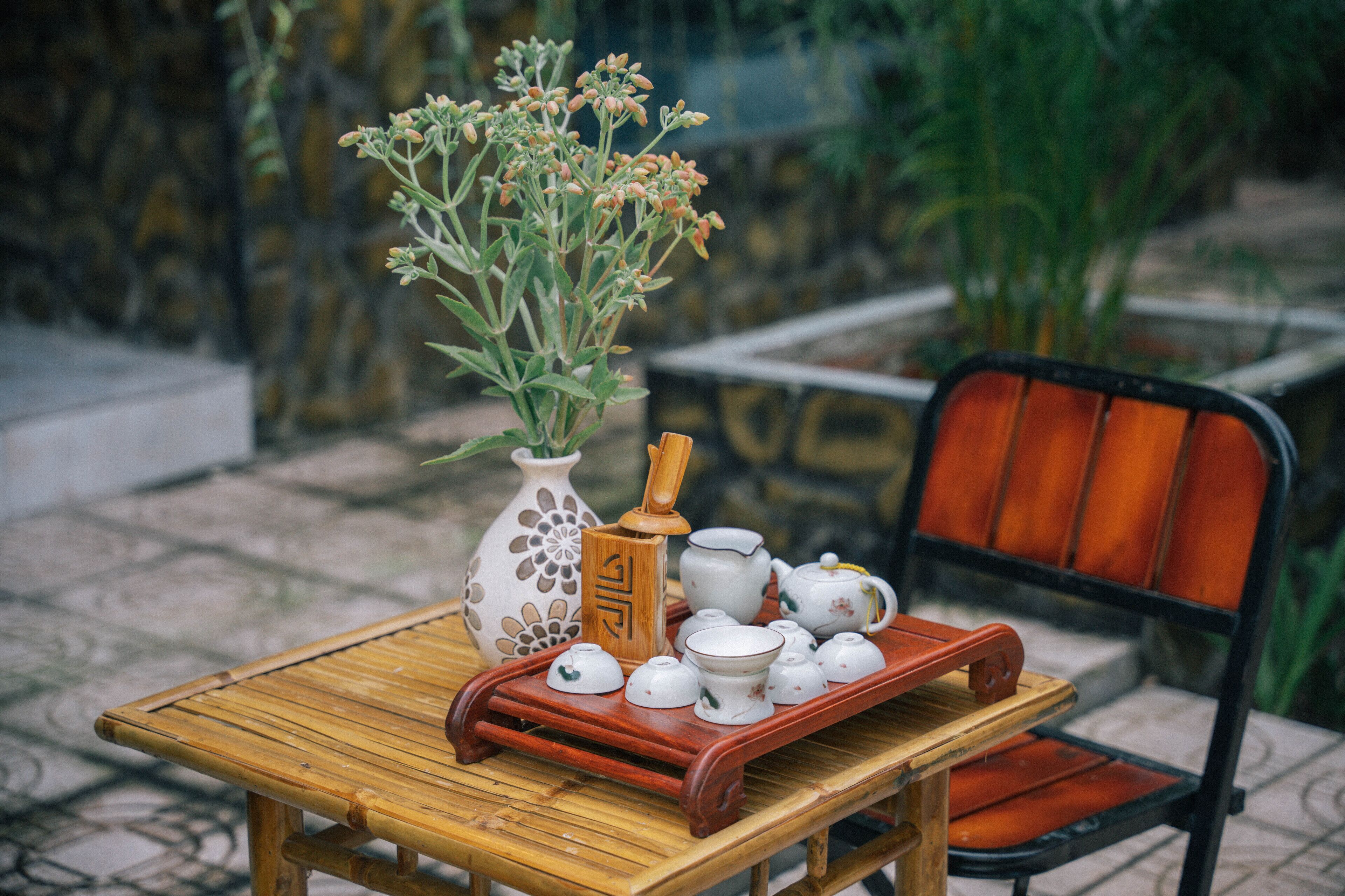 A table with a vase of flowers, a wooden tray with teapots, and a chair.