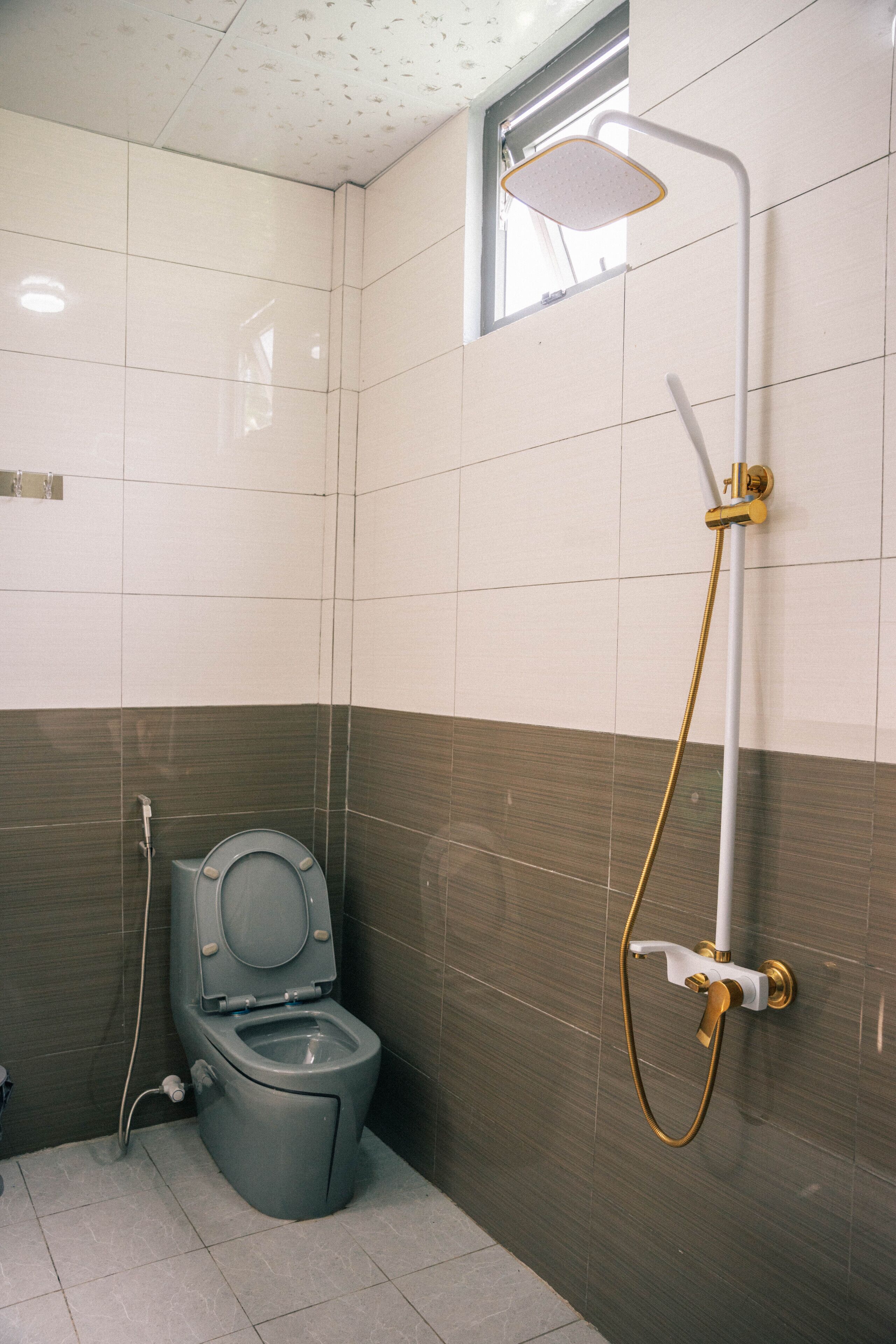 Et badeværelse med toilet, brusebad med håndbruser og et vindue over toilettet.