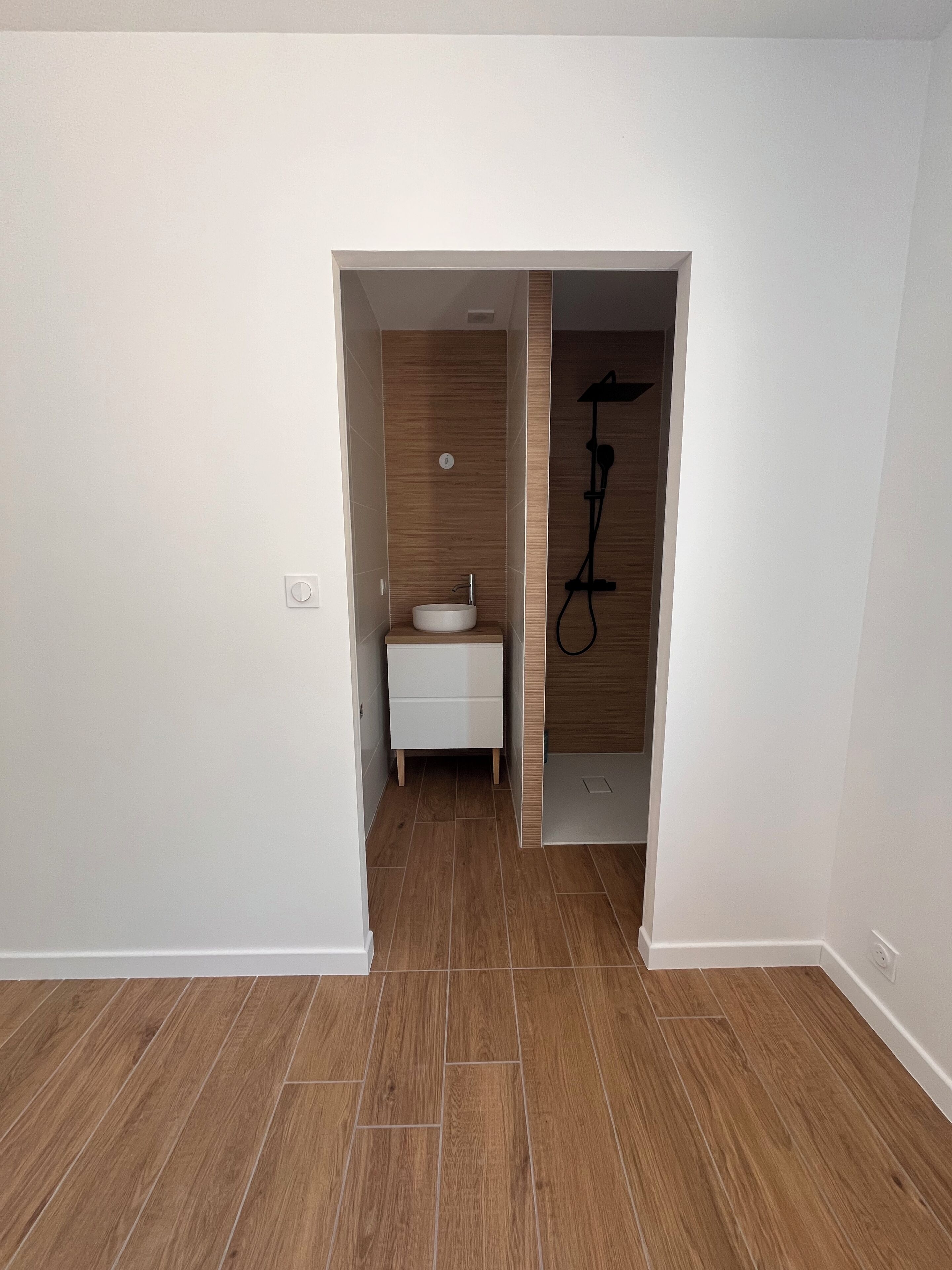 A narrow hallway leading to a bathroom with a white sink, wooden flooring, and a shower.