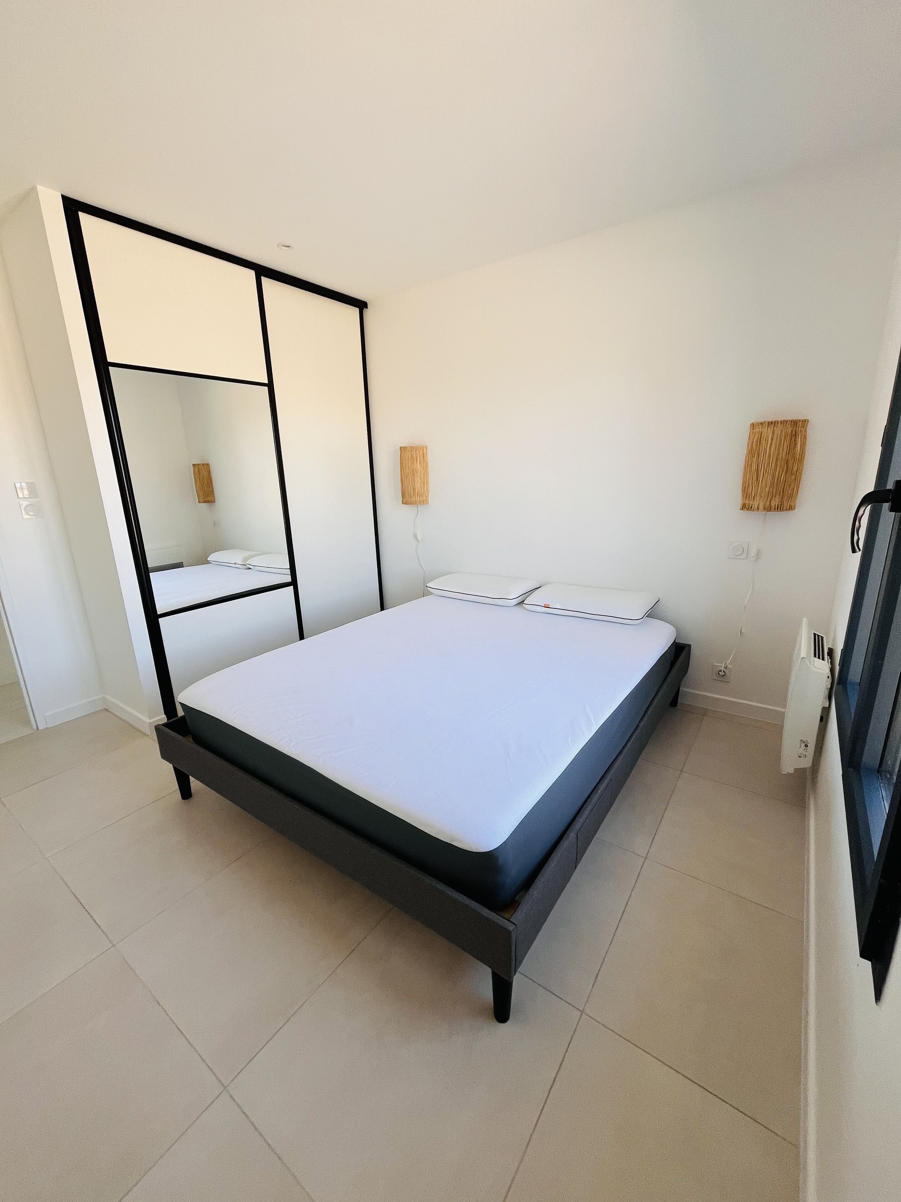 Un dormitorio moderno con una cama, un espejo grande y dos lámparas fijadas a la pared.