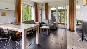 A modern living room with a sofa, a dining table with chairs, and a TV. There are large windows with curtains, and a view of greenery outside. - Bungalowpark It Wiid by Interhome (Eernewoude)