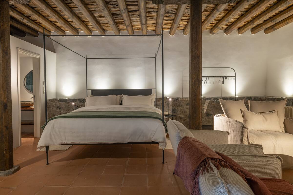 A bedroom with a four-poster bed, a stone wall, and a rustic wooden ceiling.